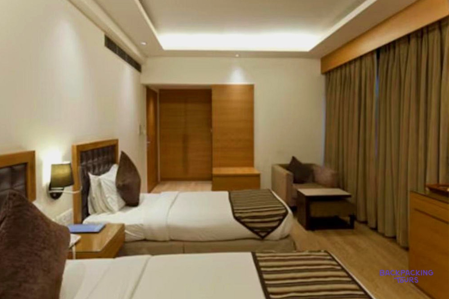 A modern hotel room features two beds with striped runners, bedside tables, a sofa, and wooden furnishings.