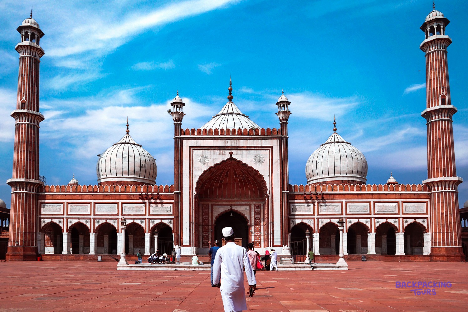 A person in white robes walks across a red courtyard towards a grand mosque with domes and minarets.