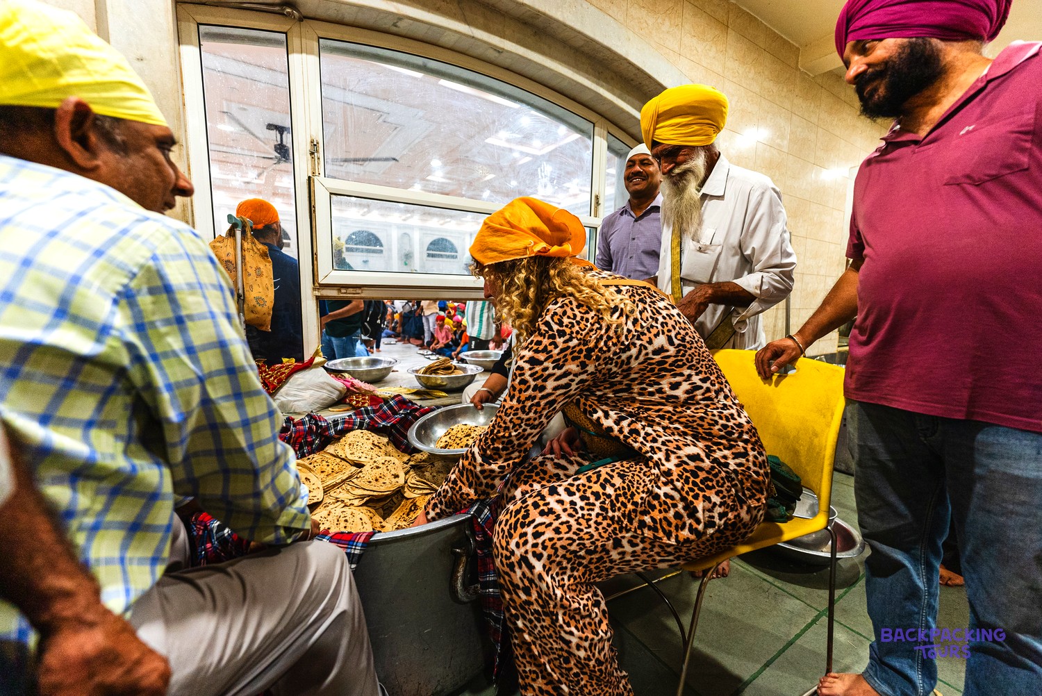 People, some in turbans, gather around flatbreads in a large container, with one woman serving.