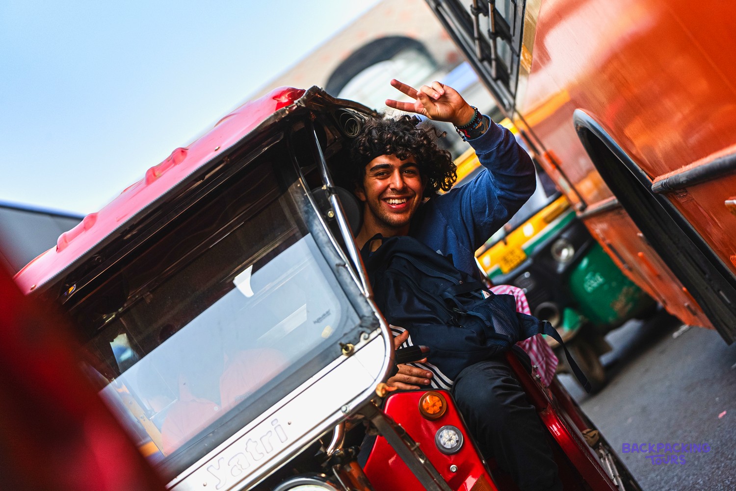 A smiling man with curly hair makes a peace sign from a red auto-rickshaw.