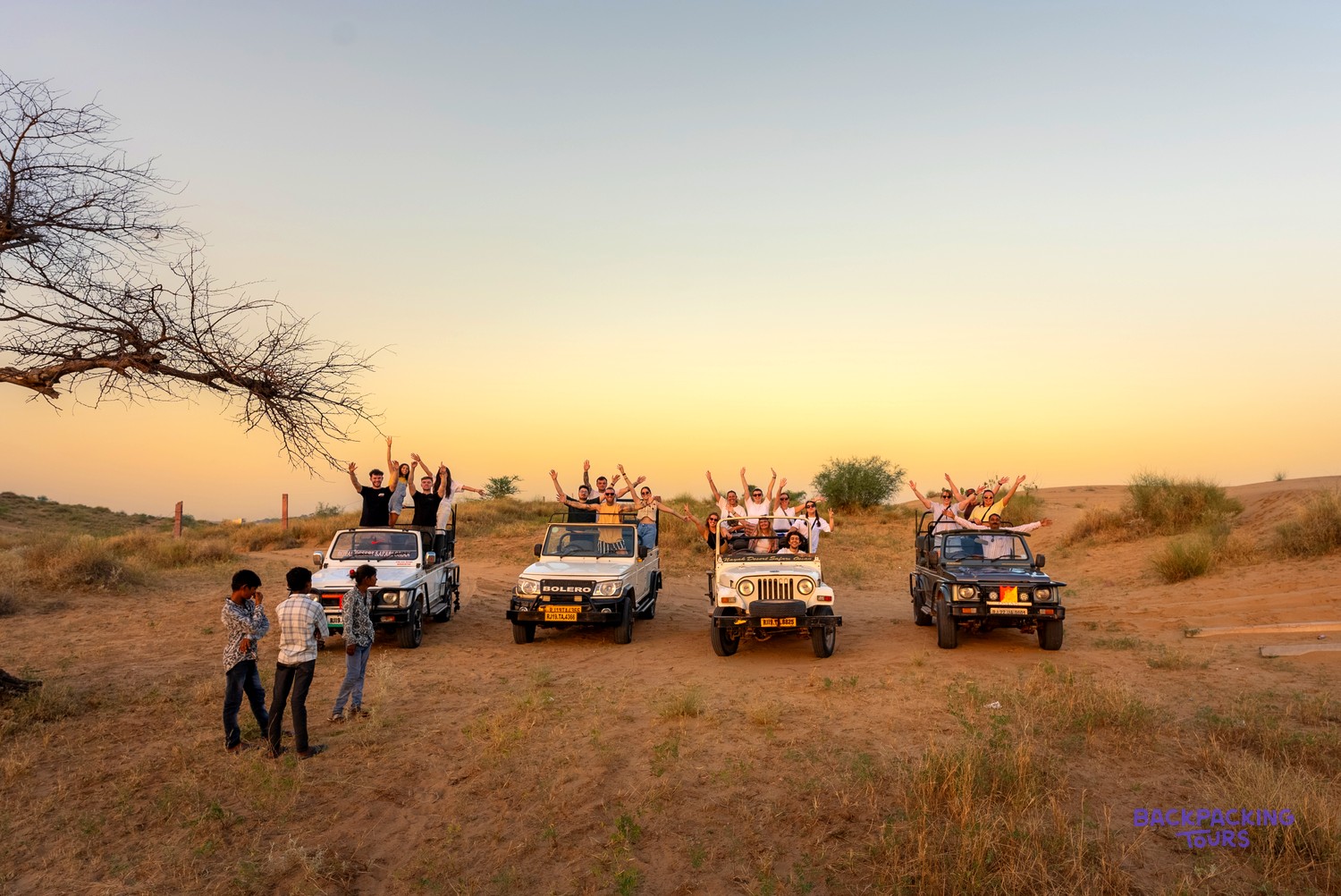 A group of people wave from open-top jeeps during a desert safari at sunset.