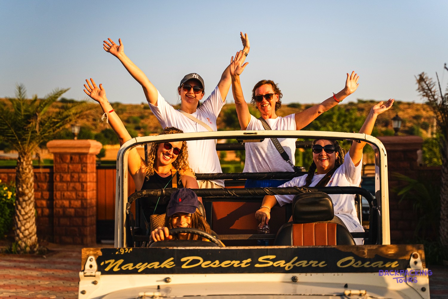 Five women smiling and waving from an open-top jeep during a desert safari.