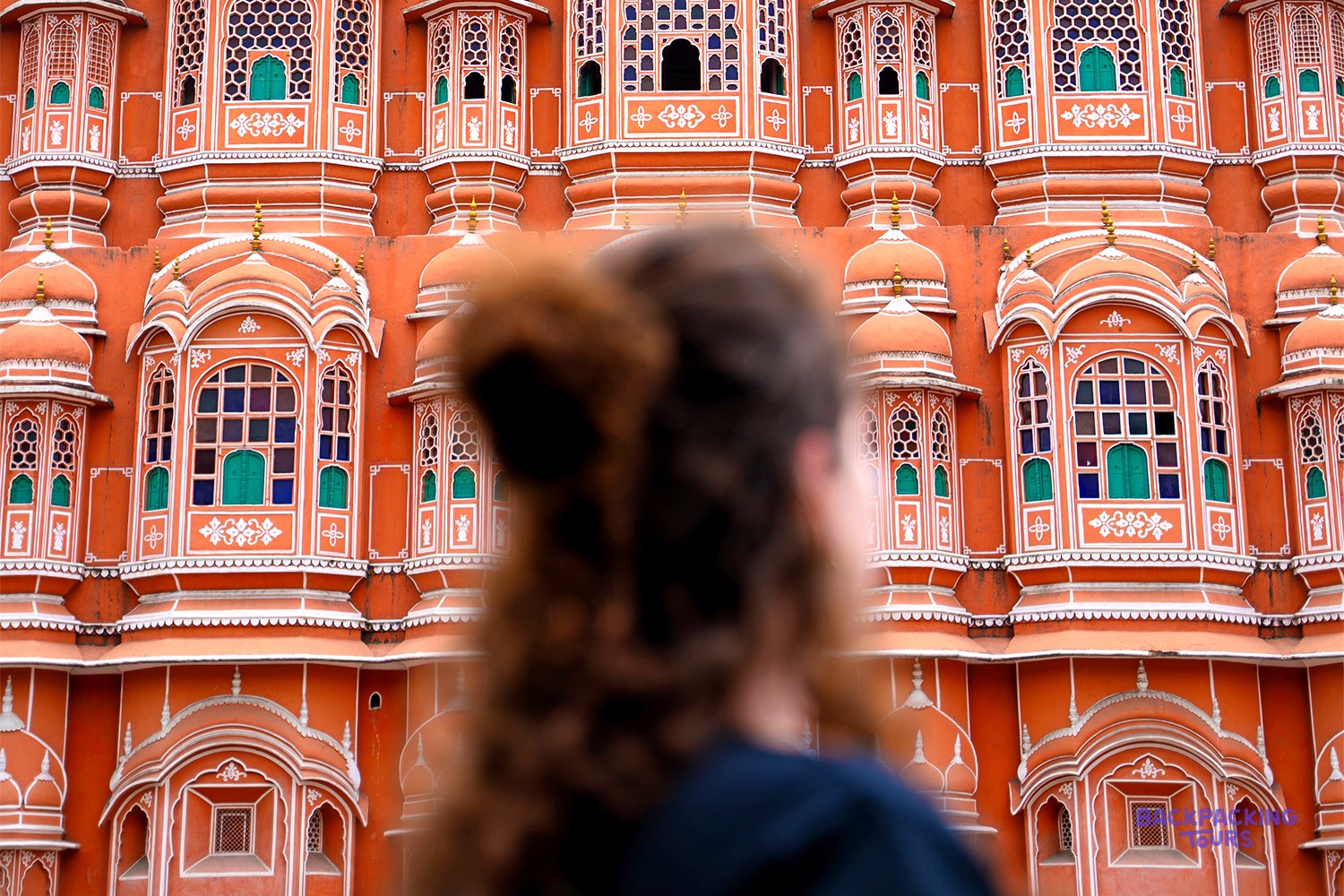 A traveller, blurred in the foreground, looks up at an ornate orange building with many small windows.