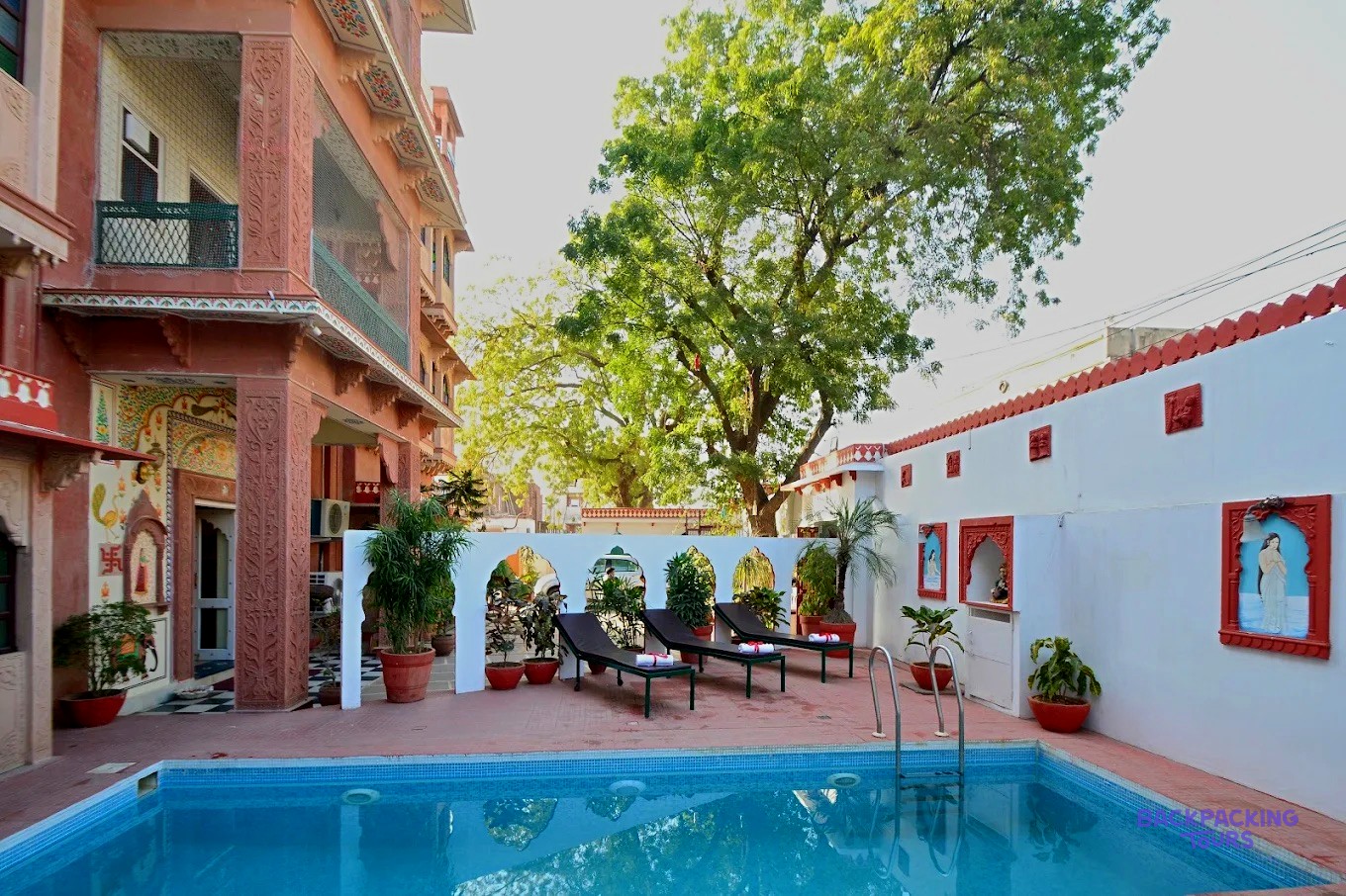 A swimming pool with sun loungers is surrounded by a decorative white wall and a multi-storey building.