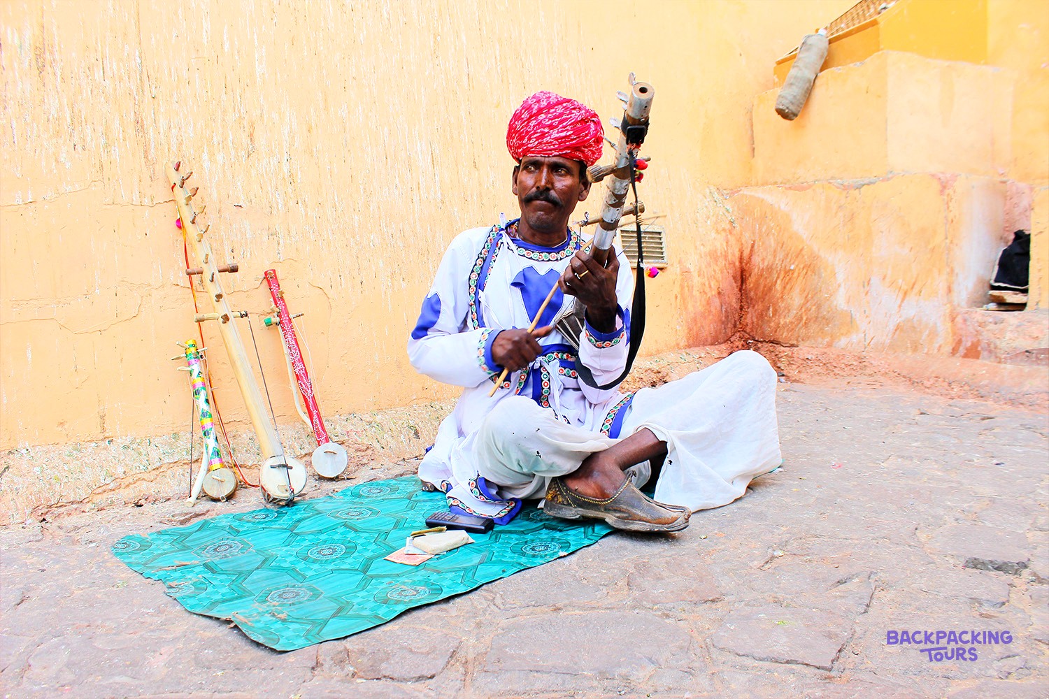 A musician in a red turban and white tunic plays a stringed instrument, with other instruments nearby.