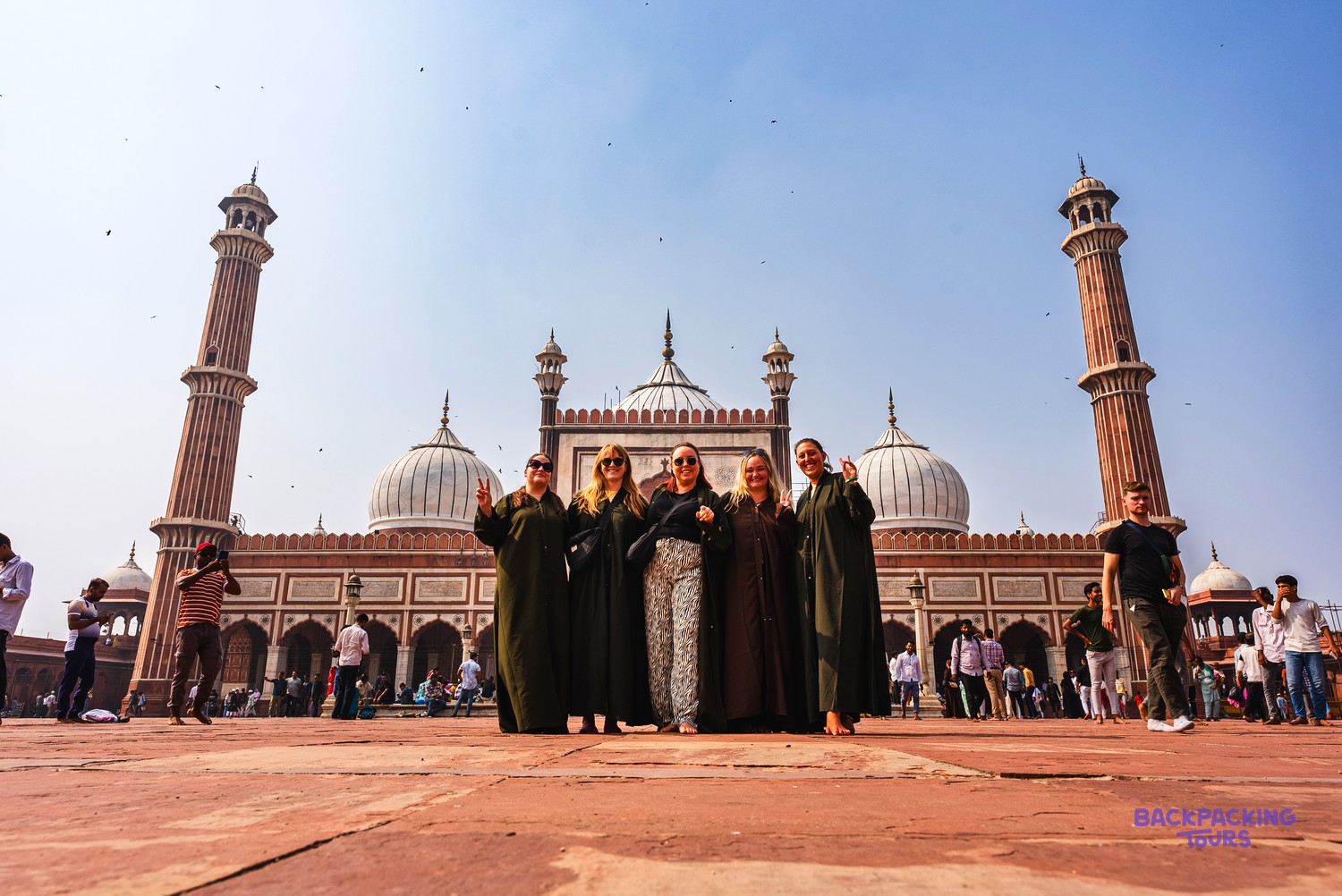 Five women pose on a large red courtyard in front of a grand mosque with domes and minarets.