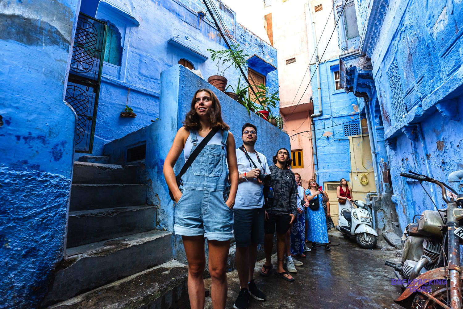 A group of travellers stands in a narrow lane between tall, blue-painted buildings.