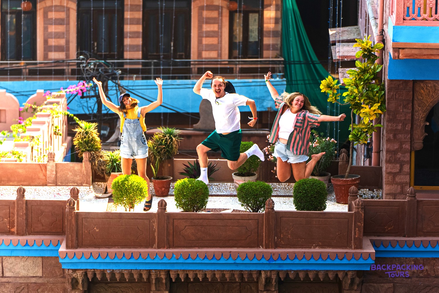 Three young people jump high on a colourful rooftop terrace with potted plants and buildings.