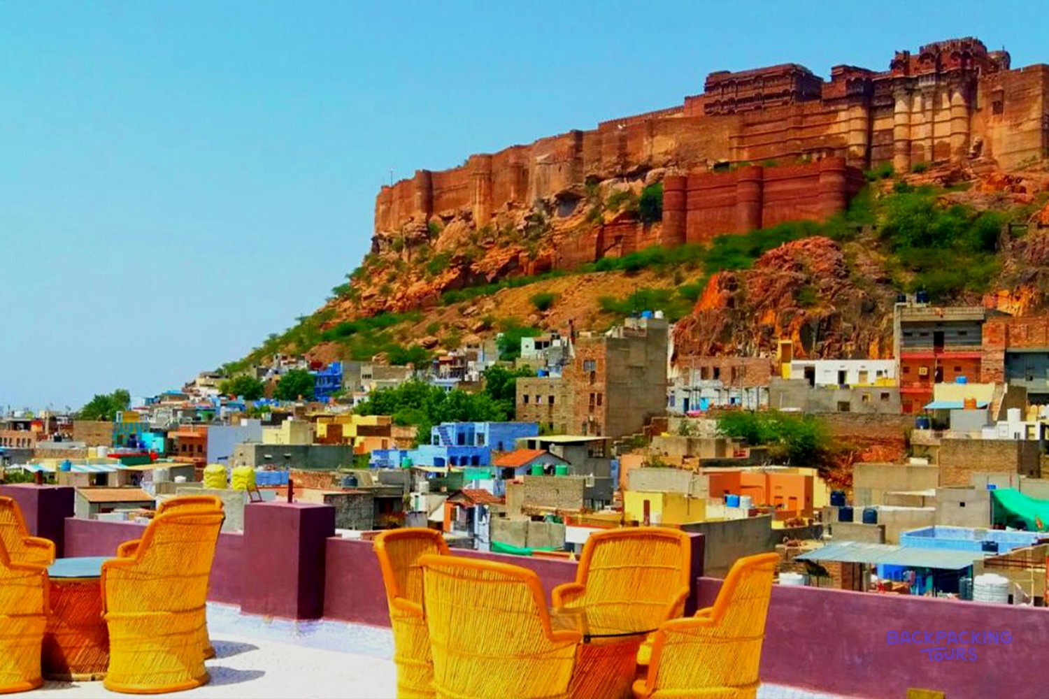A rooftop terrace with yellow chairs provides views of a colourful city and a large fort.