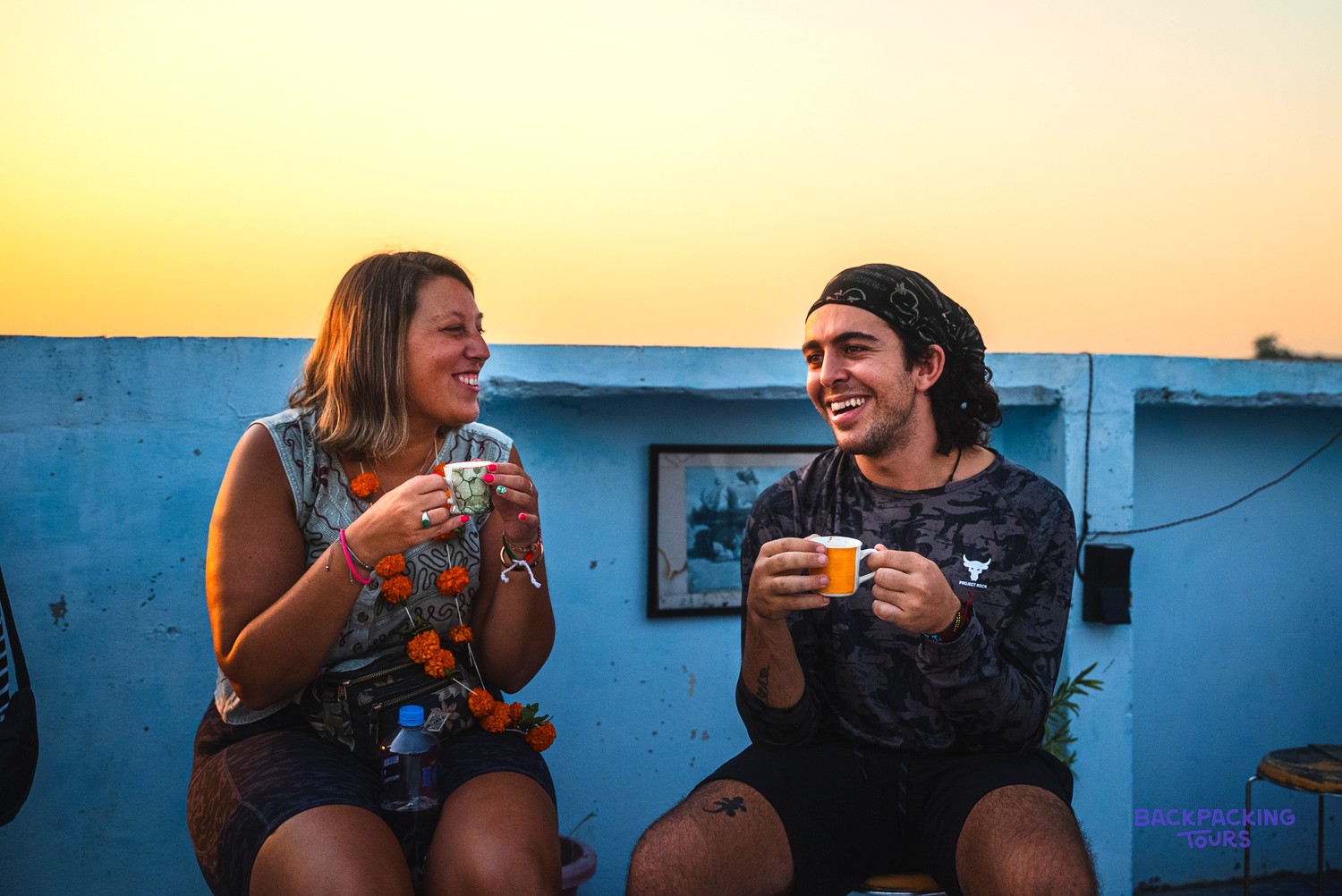 Two smiling people on a blue rooftop at sunset, holding small cups.