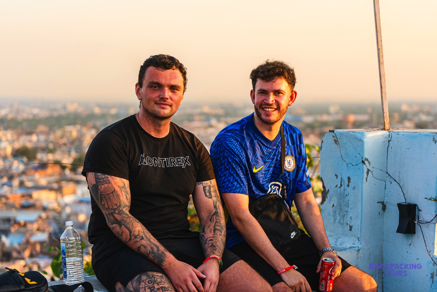 Two men smile at the camera from a rooftop overlooking a city at sunset.