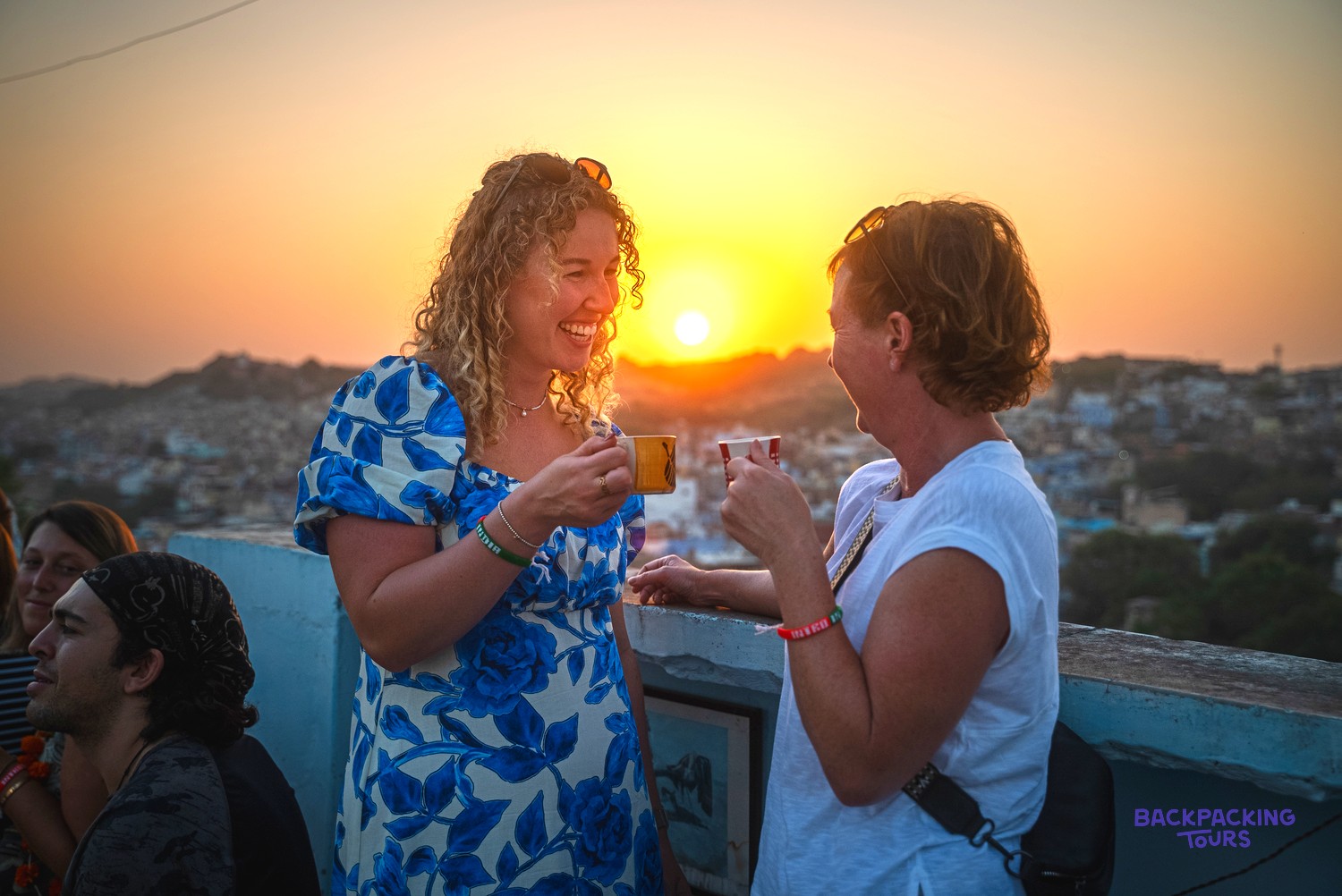 Two women smile and chat, holding cups on a rooftop overlooking a city at sunset.
