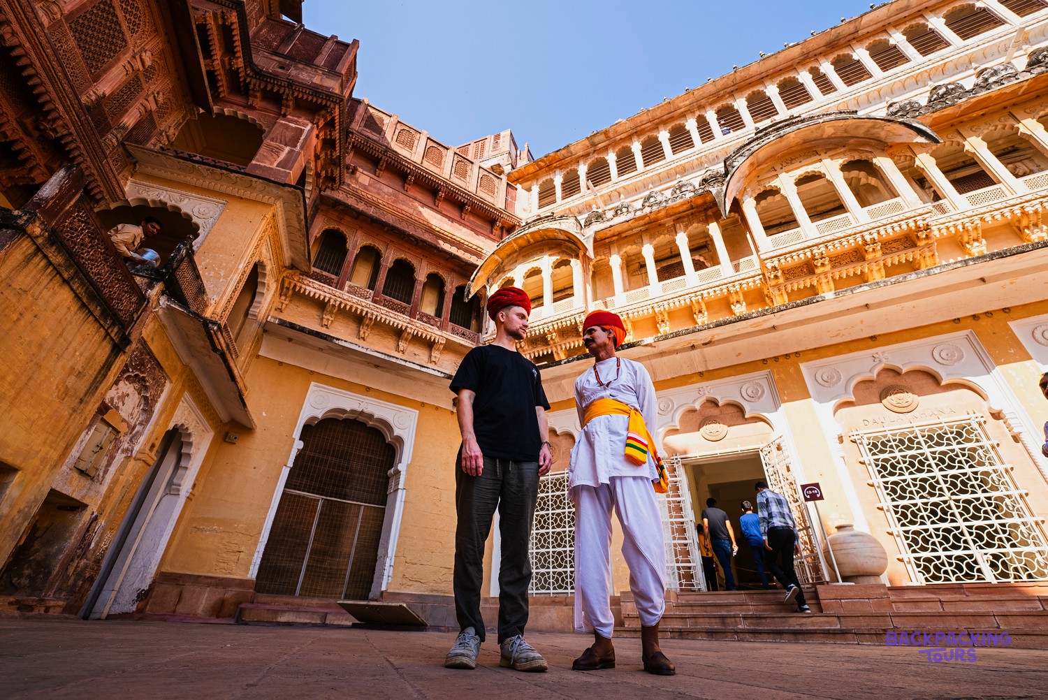 Two men stand in a grand courtyard surrounded by ornate, multi-coloured stone buildings with arched windows and balconies.