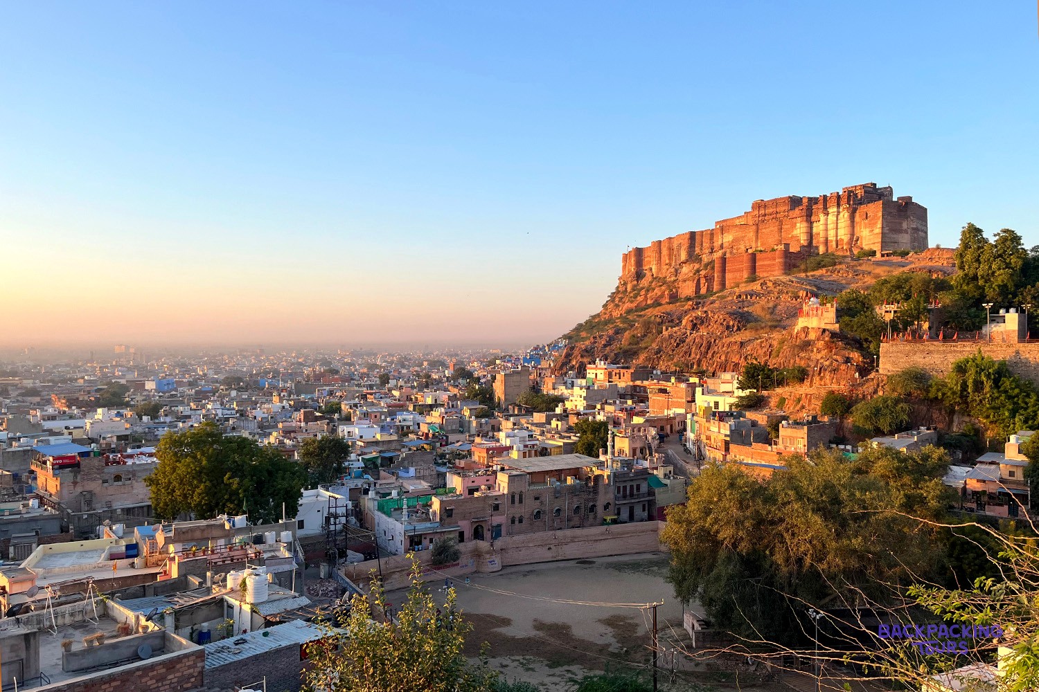 A large, ancient fort sits on a rocky hill overlooking a sprawling city with many buildings under a clear sky.