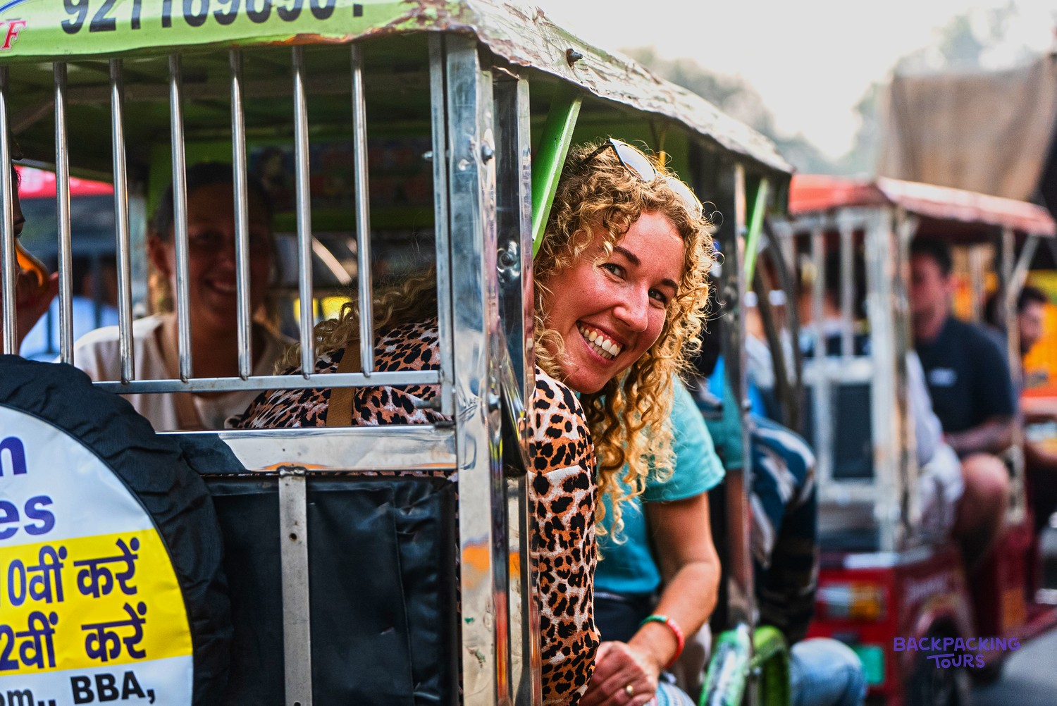 A smiling woman with curly blonde hair looks out from a caged rickshaw, with other people travelling nearby.