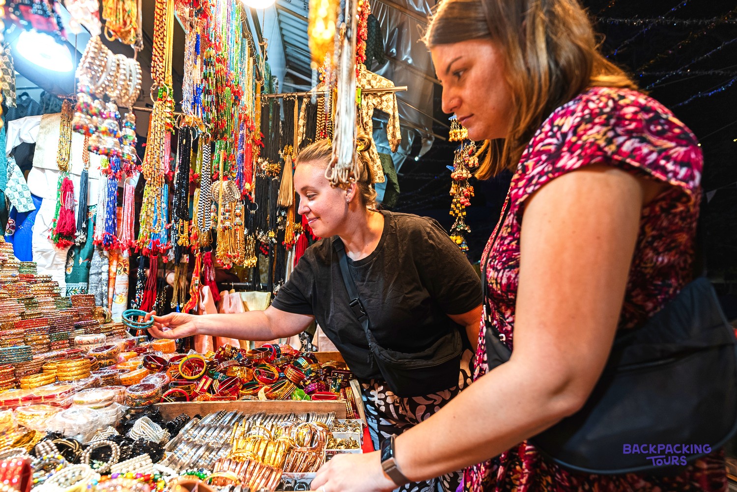 Two women browse a market stall filled with colourful bangles and necklaces, one holding a blue bangle.