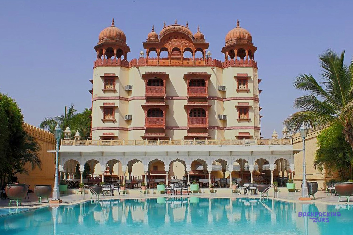 An ornate, multi-domed building with balconies overlooks a turquoise swimming pool and outdoor seating.