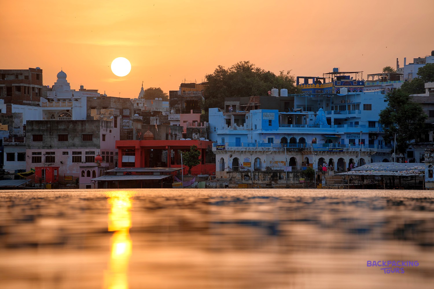 Colourful buildings line a lake, reflecting the bright orange sunset in the water.