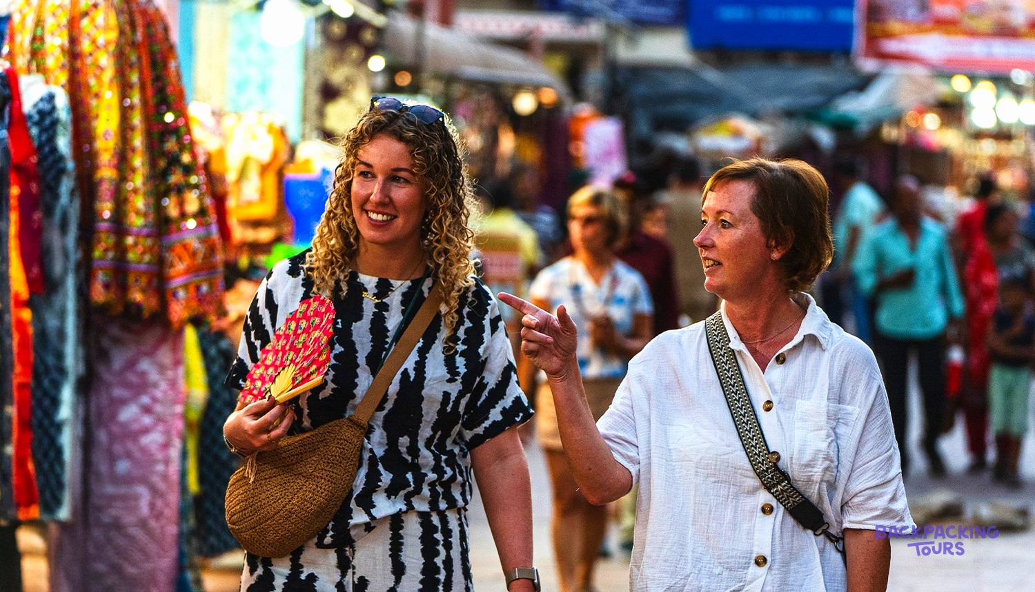 Two women walk through a bustling market street, one smiling and holding a red fan.