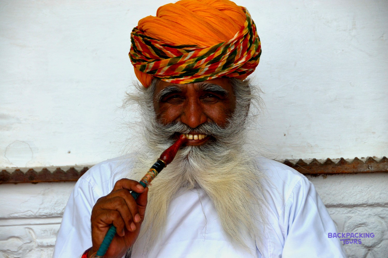 A man with a prominent white beard and an orange, patterned turban holds a pipe.