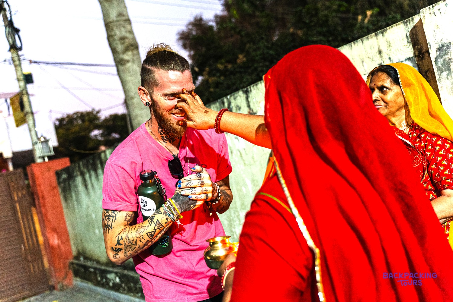 A woman in a red sari applies a colour mark to a man's forehead, while another woman watches.