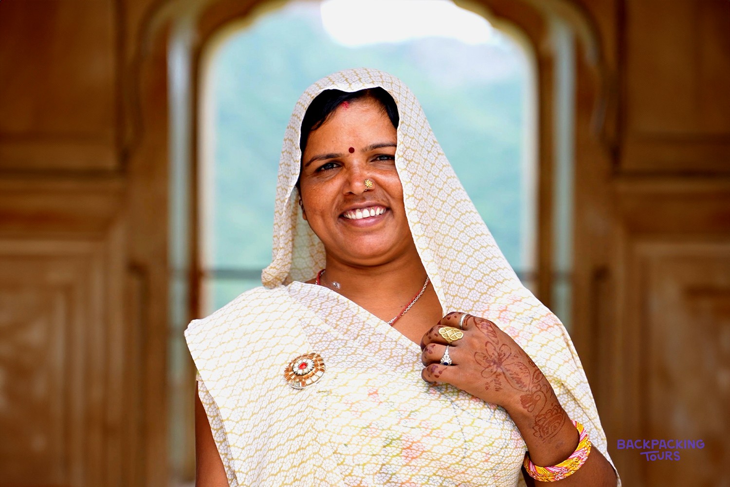 A smiling woman in a patterned white head covering, sari, jewellery, and henna stands before an arched doorway.