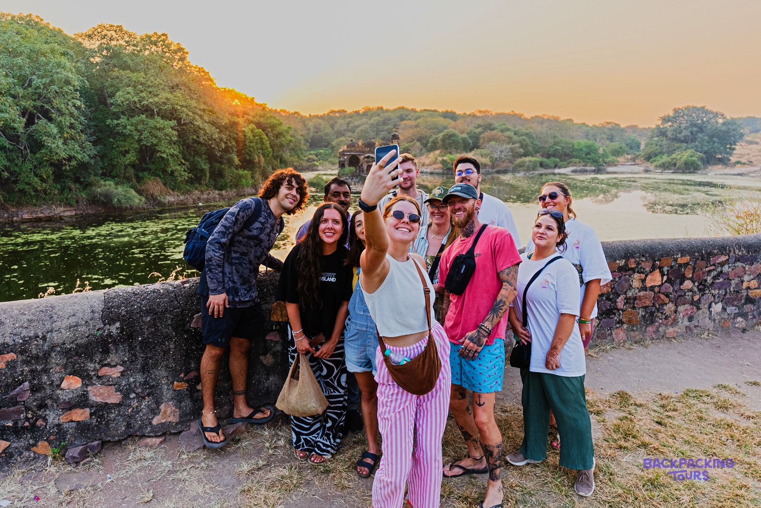 A group of travellers taking a selfie on a stone wall beside a lake at sunset.