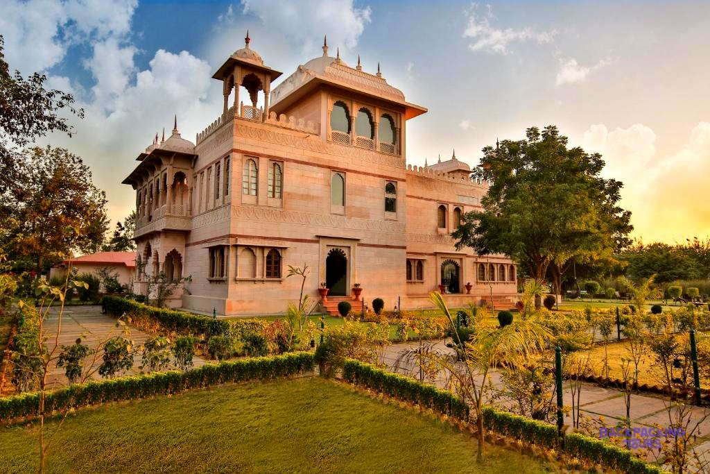 An ornate, light-coloured building with domes and arched windows stands in a lush garden at sunset.