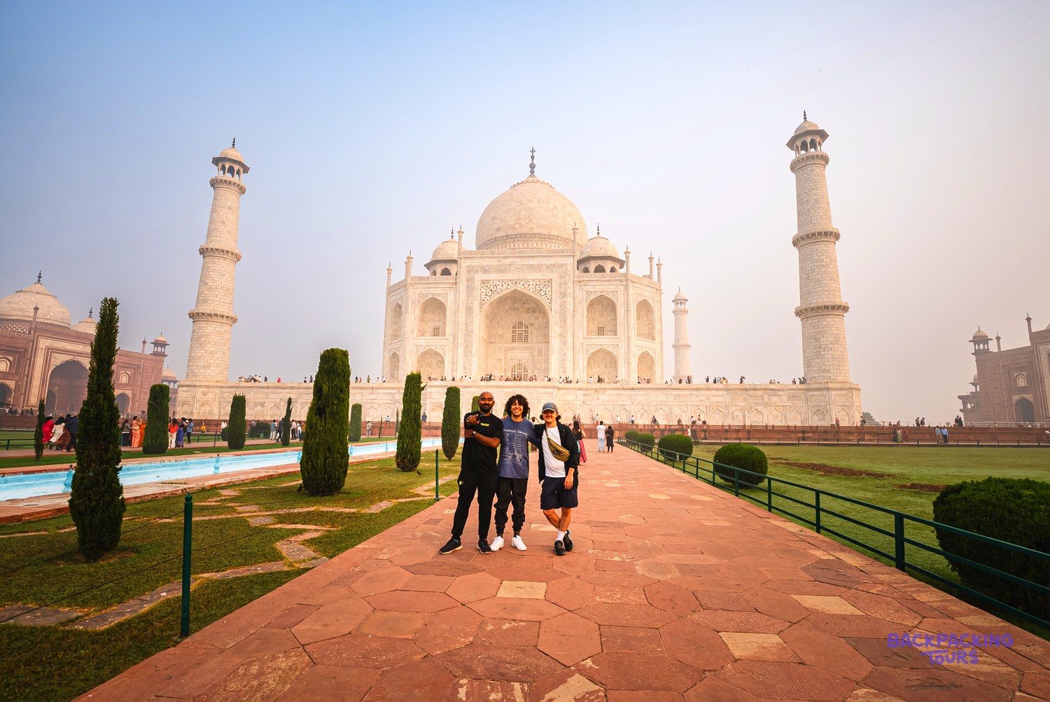 Three people pose on a red path in front of the white marble Taj Mahal at sunrise.