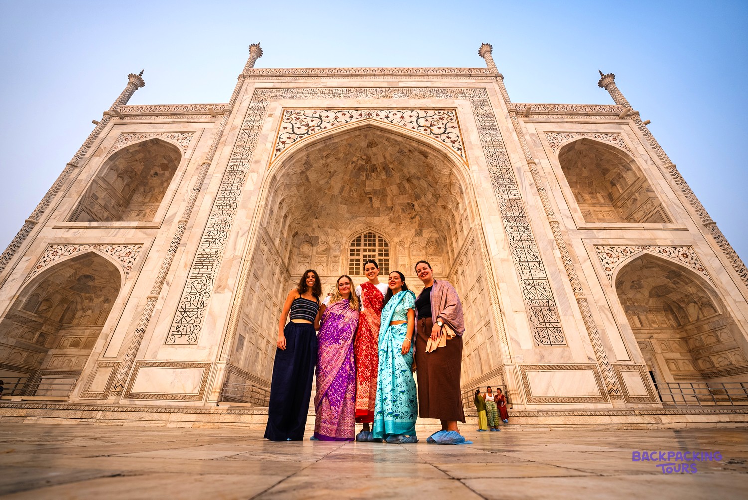 Five women pose smiling in front of the majestic white marble Taj Mahal.