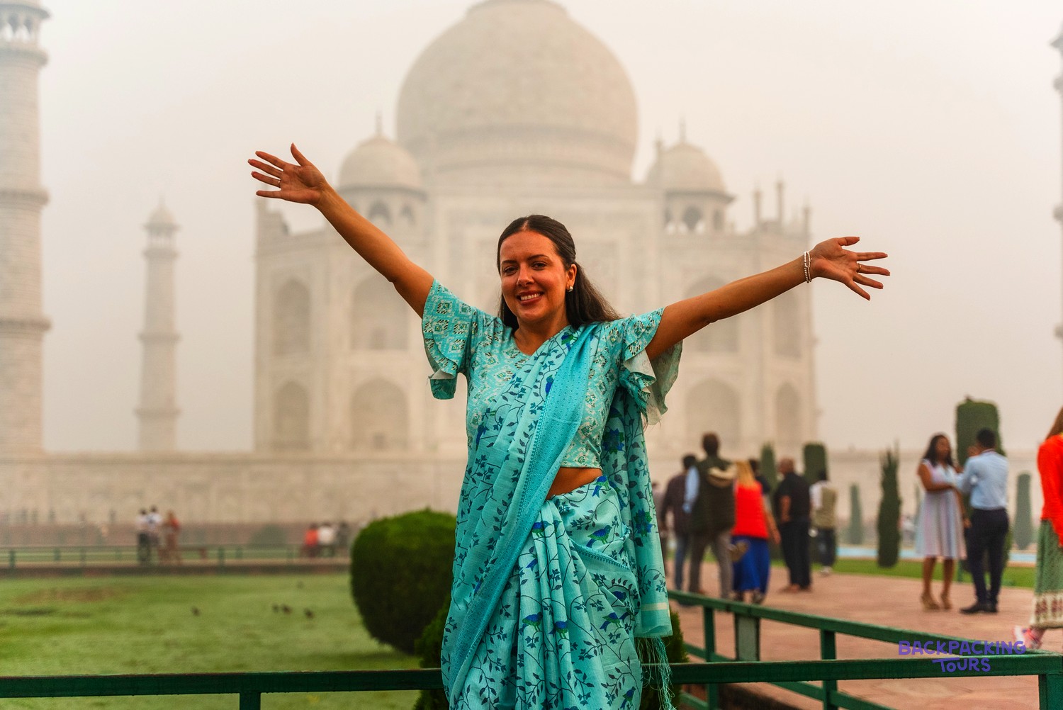 A woman in a blue patterned sari stands with arms outstretched before a large white domed building.