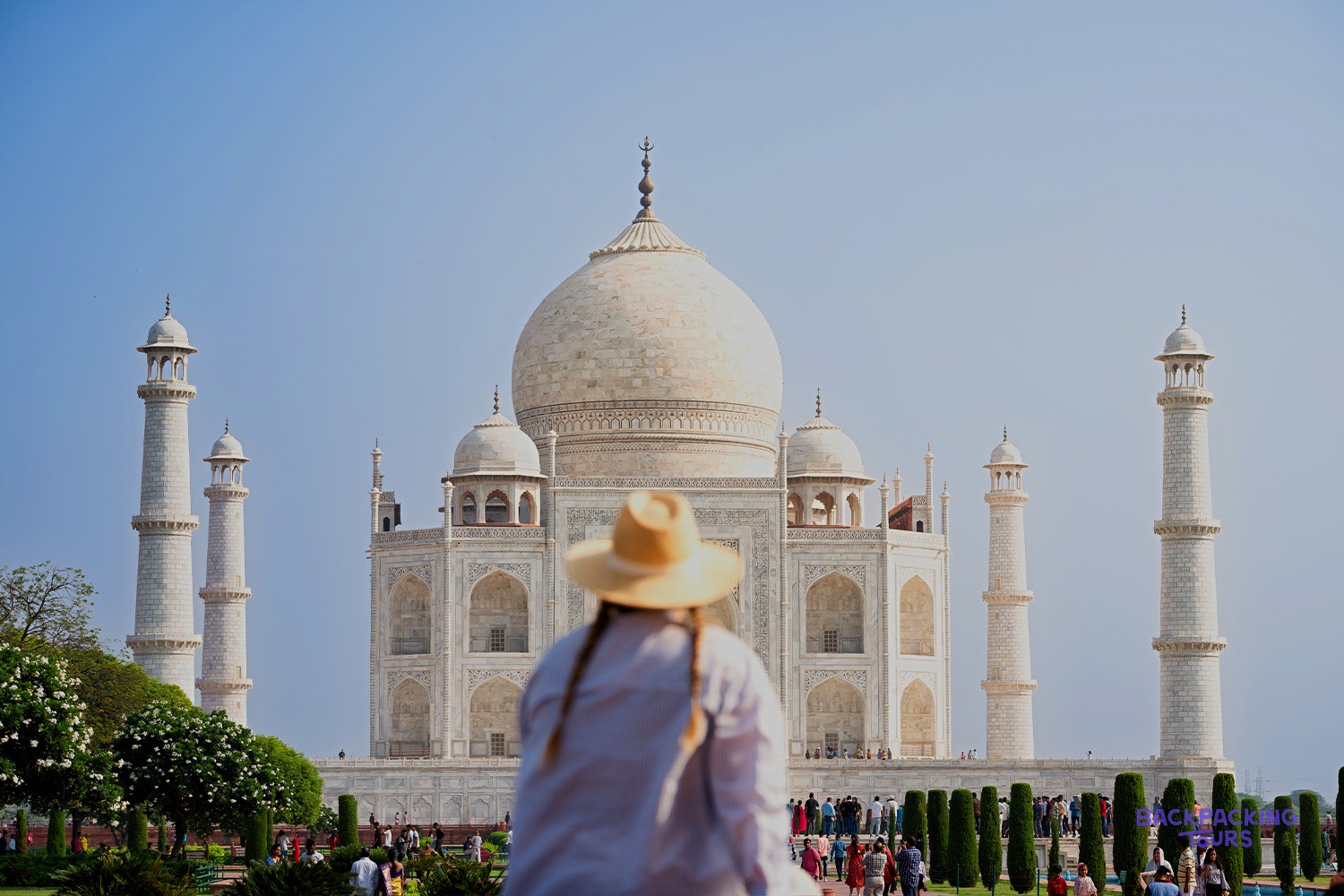 A person in a straw hat views a grand white marble building with a large dome and four tall minarets.