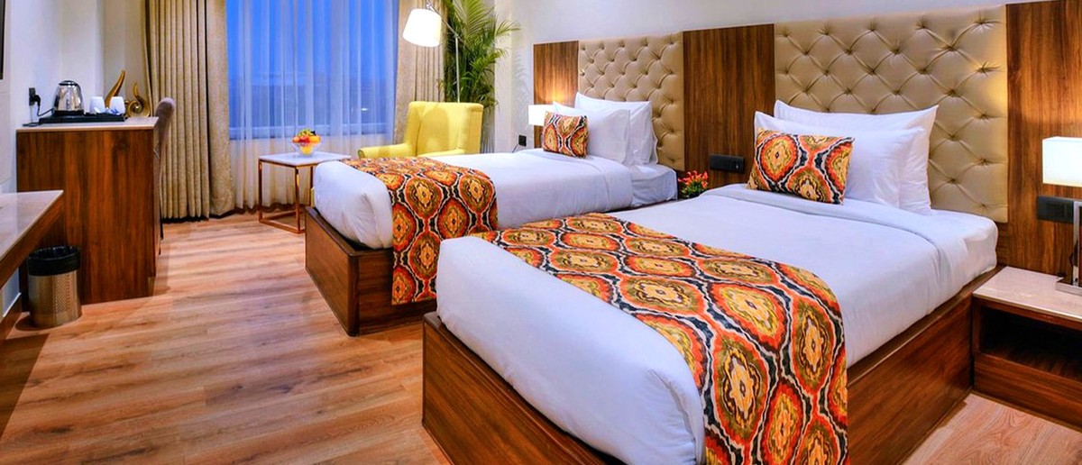 A modern hotel room features two beds with colourful patterned runners, a yellow armchair, and a window.
