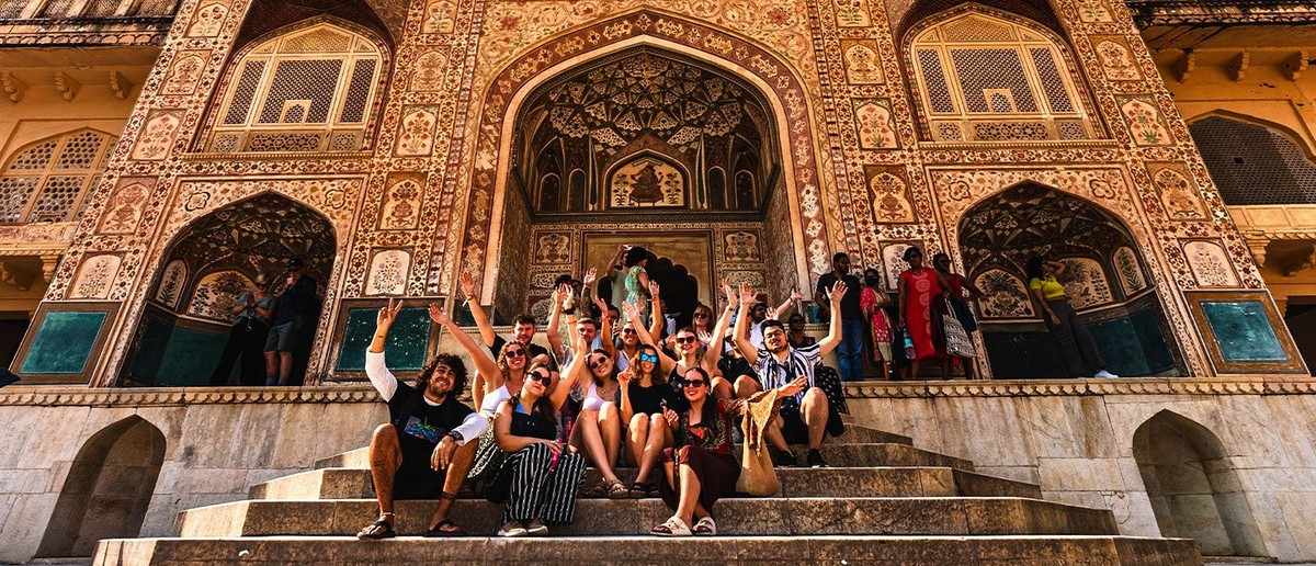 A group of travellers sits on stone steps, smiling and waving in front of an ornately decorated building.