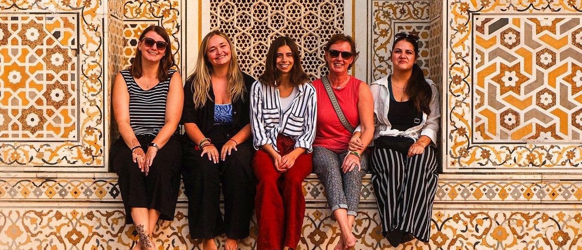 Five women sit barefoot against an ornate wall decorated with yellow and grey geometric patterns.