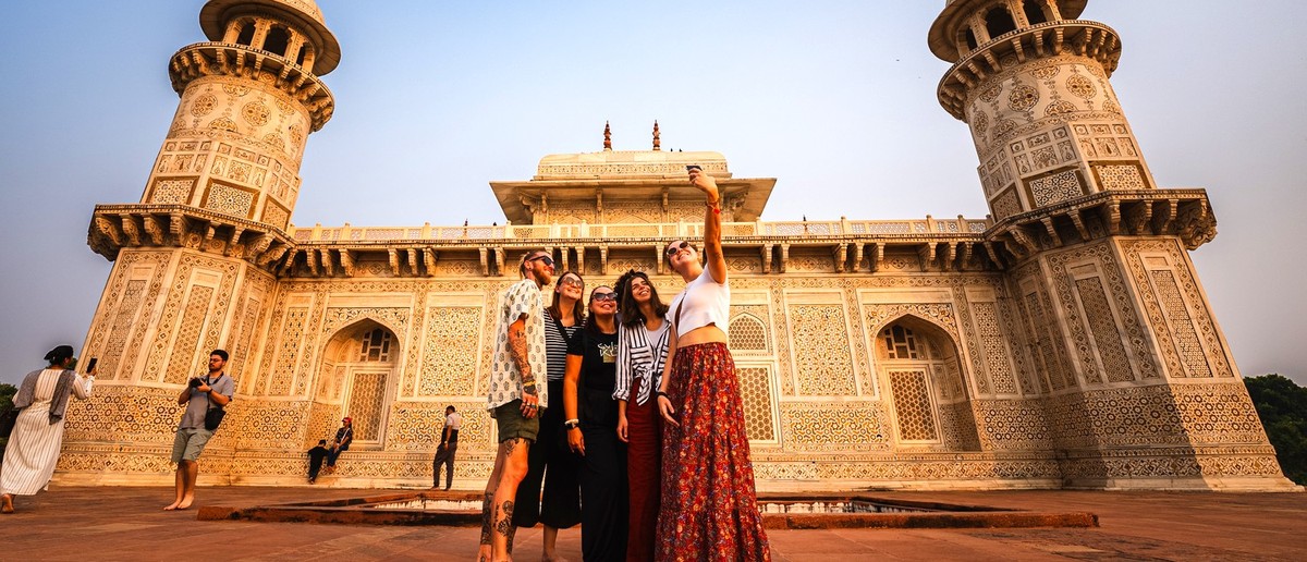 Five young people take a selfie in front of an intricately carved white marble building with two minarets.