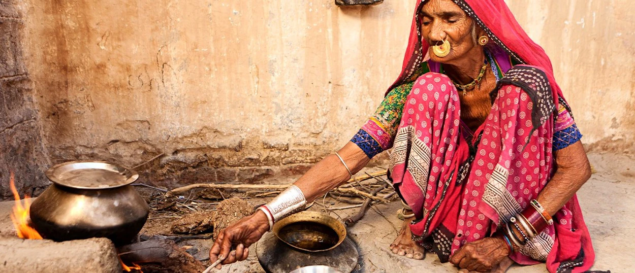 An elderly woman in colourful traditional dress squats, stirring a cooking pot over an open fire.