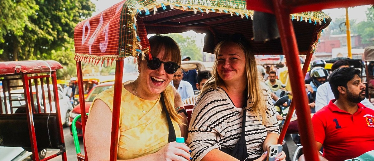 Two women are smiling and laughing while travelling in a red rickshaw on a busy street.