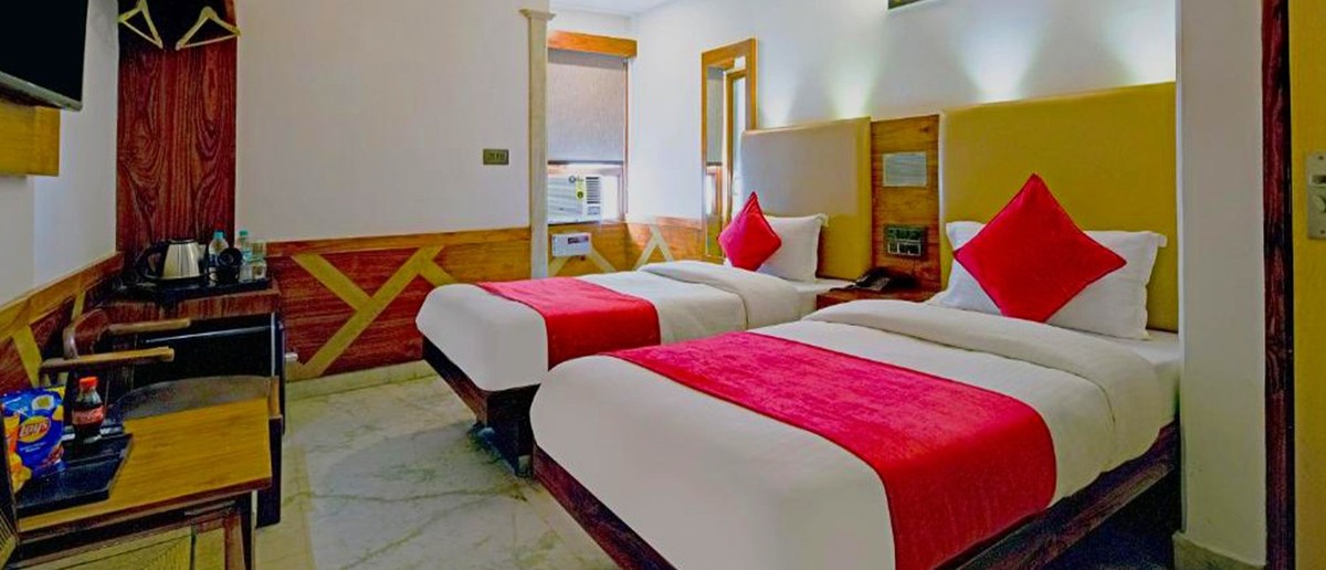 A bright hotel room features two single beds with red throws, a ceiling fan, and a small desk.