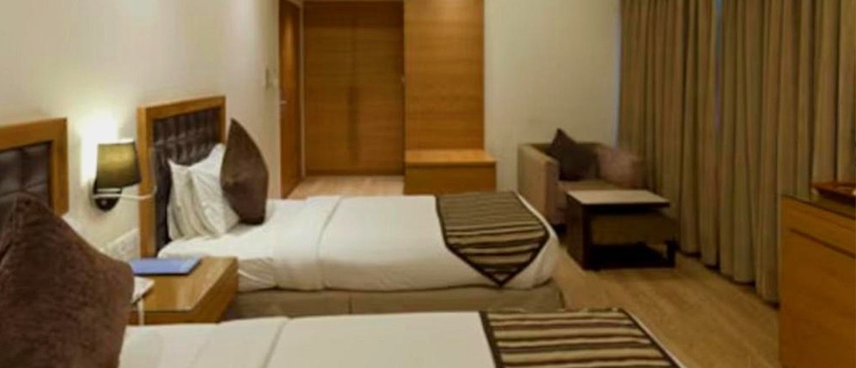 A modern hotel room features two beds with striped runners, bedside tables, a sofa, and wooden furnishings.