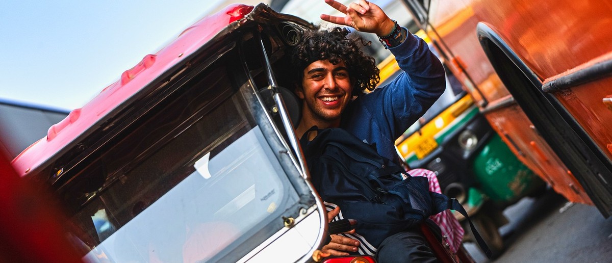 A smiling man with curly hair makes a peace sign from a red auto-rickshaw.