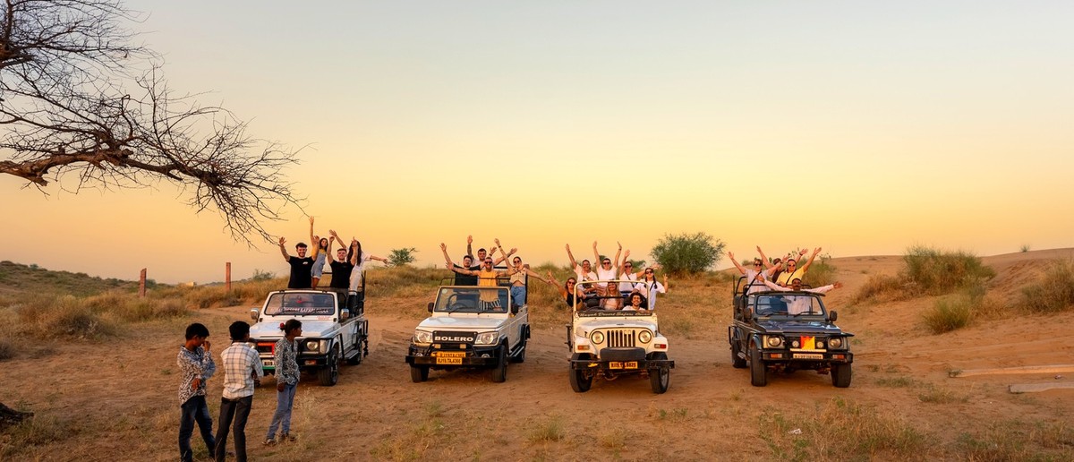 A group of people wave from open-top jeeps during a desert safari at sunset.
