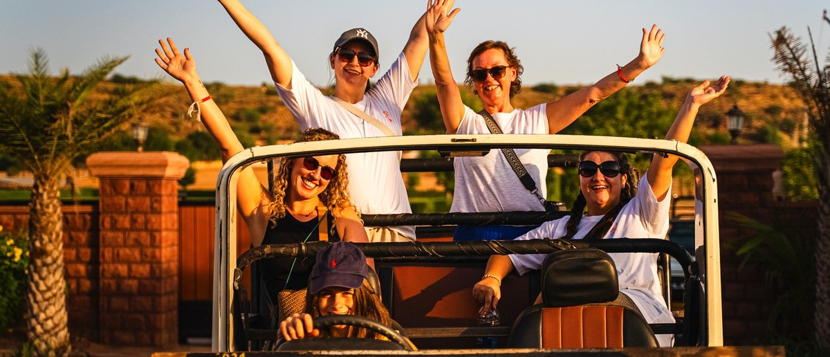 Five women smiling and waving from an open-top jeep during a desert safari.