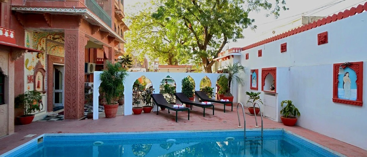 A swimming pool with sun loungers is surrounded by a decorative white wall and a multi-storey building.