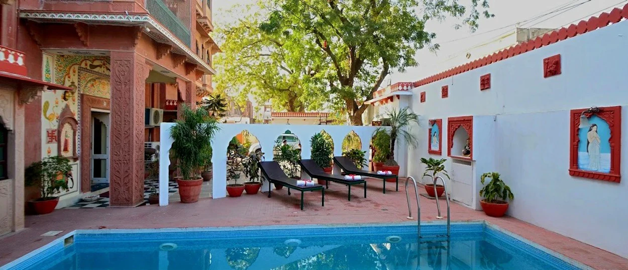 A swimming pool with sun loungers is surrounded by a decorative white wall and a multi-storey building.