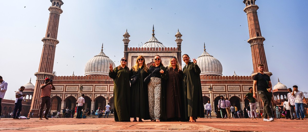 Five women pose on a large red courtyard in front of a grand mosque with domes and minarets.