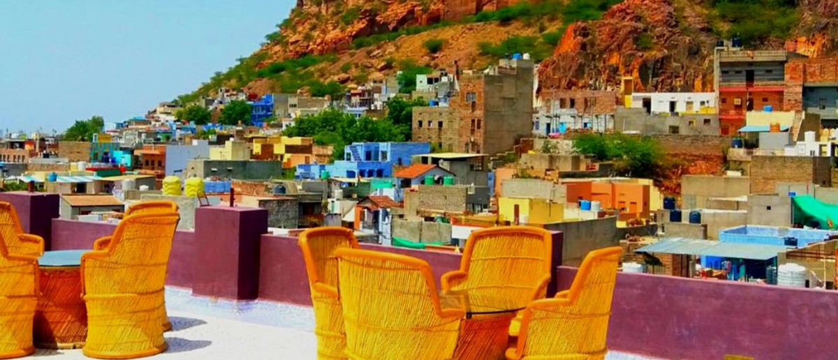 A rooftop terrace with yellow chairs provides views of a colourful city and a large fort.