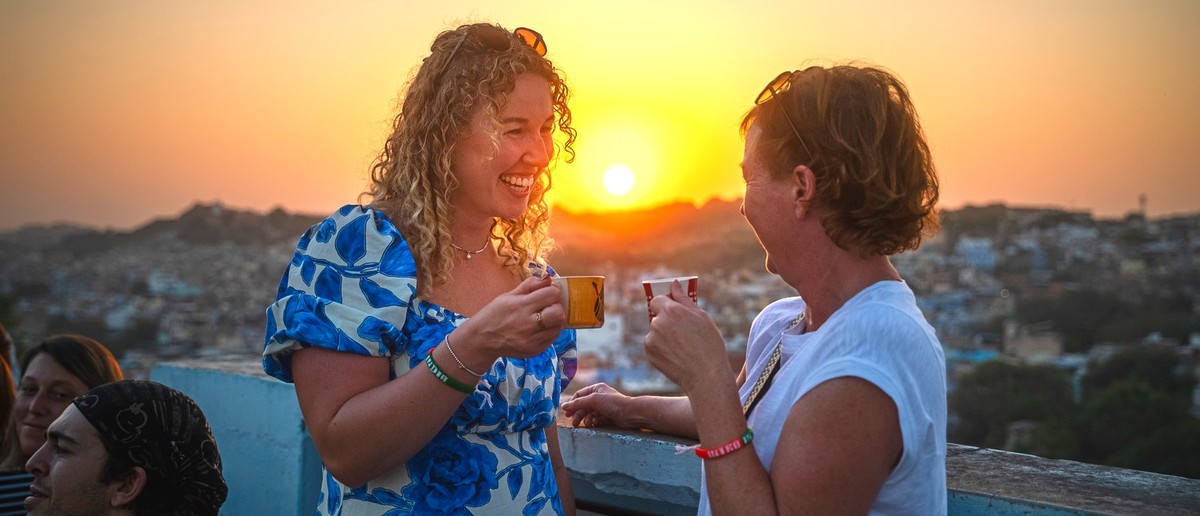 Two women smile and chat, holding cups on a rooftop overlooking a city at sunset.