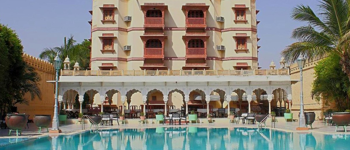 An ornate, multi-domed building with balconies overlooks a turquoise swimming pool and outdoor seating.