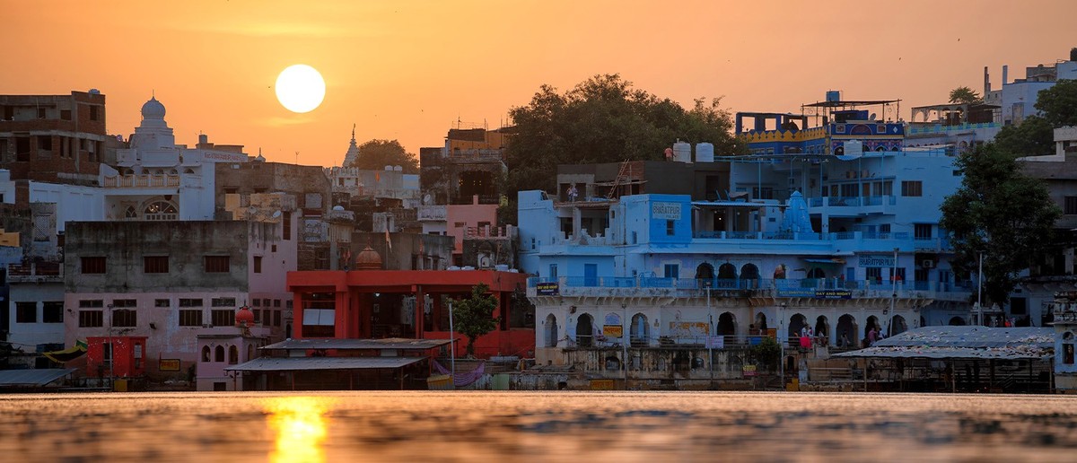 Colourful buildings line a lake, reflecting the bright orange sunset in the water.